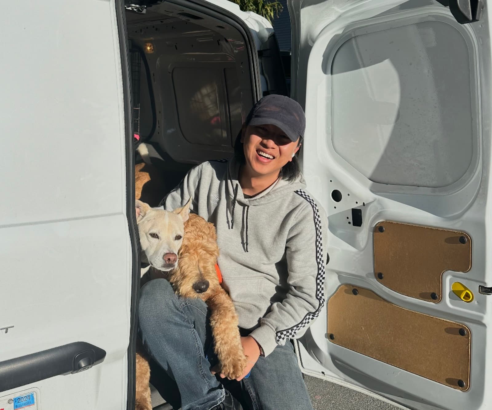 A staff member sitting in the open door of the white Tefani shuttle van, smiling and holding two dogs in the sunlight